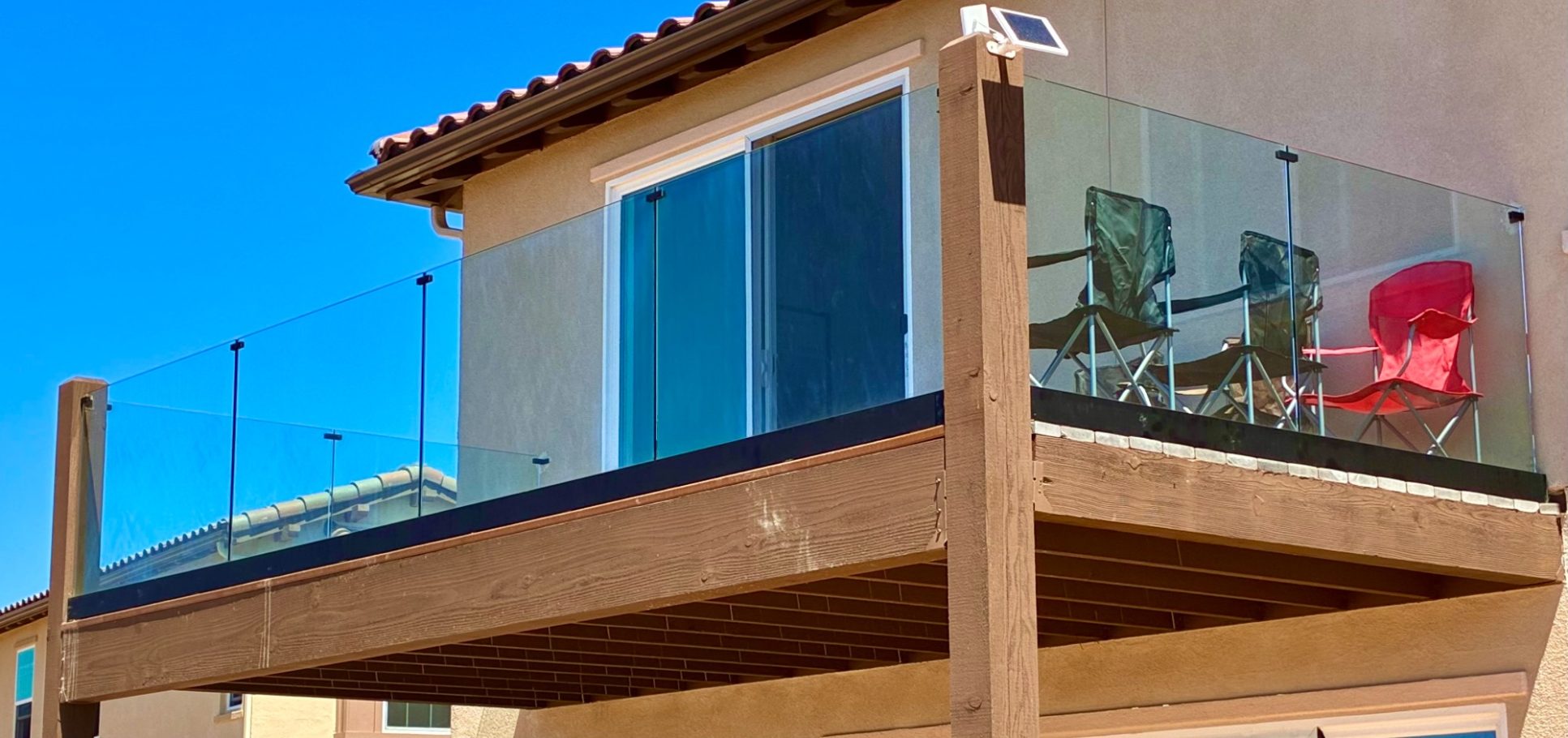 Second Story Frameless Balcony Railing Modern wooden deck with glass railing and two chairs, under a clear blue sky.